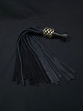 Black leather flogger with a braided gold accent and a ball handle meant to sit between the index and middle finger with the flat top pressed against the palm. There is a triskellion engraved on the top of the handle. Displayed on a black fabric background.