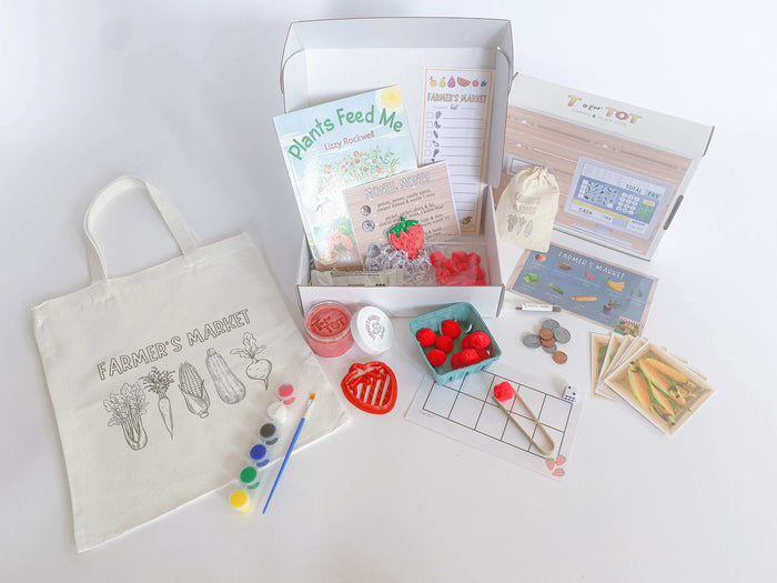 Farmer's Market play and learn kit for kids. Features strawberry-scented playdough with custom strawberry cutter, 'Plants Feed Me' book, and a canvas tote bag with paint for custom decoration. Includes play money with a cash register sticker, a tens frame math game, market signs, and carrot seeds for planting. Perfect for imaginative play and learning at home.