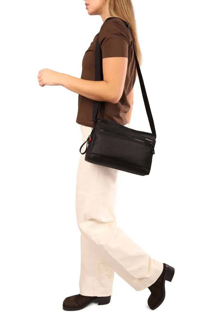 Hedgren EYE NEO Medium Crossbody Bag with RFID Pocket in Green Milieu, worn crossbody at the hip by a woman walking in a brown tee and cream trousers