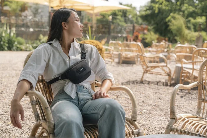 Pacsafe Go Anti-Theft Micro Sling in Jet Black, worn across the chest by a woman sitting in a cane chair at an outdoor bar