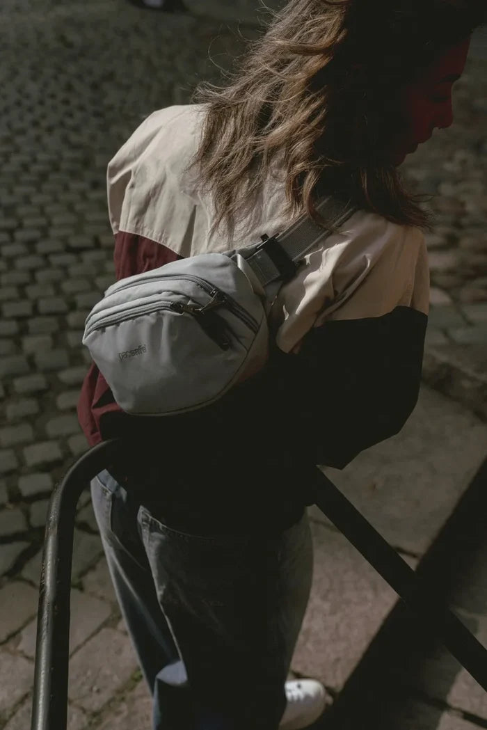 Pacsafe V Anti-Theft Hip Pack in Beige, worn crossbody over a jacket on a set of stone steps